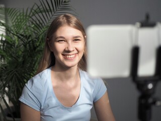 Girl blogger influencer smiling face, talking with followers, live streaming, recording video blog looking to smartphone on tripod. Video call to friends, parents. Selected Focus