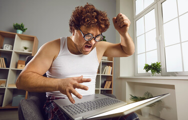 Funny angry curly guy having problem with his computer at home. Furious crazy nerdy young man...