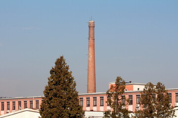 Fototapeta premium Red bricks tall industrial chimney with metal stairs mounted on side rising high above elongated old factory building and trees on warm sunny winter day