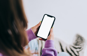 Woman holding a smartphone with a white screen mock up, resting on a sofa in living room at home.