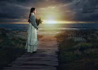 Fototapeta premium Young victorian girl holding dried flowers in white dress and blue striped blouse on a boardwalk at the coast at sunset.
