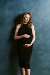 Fototapeta na wymiar Smiling pregnant brunette young woman with long curly hair, in elegant black midi dress. Fashion style studio photoshoot of pregnancy. Between the second and third trimesters of pregnancy