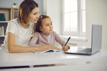 Small positive girl and her mother sitting together in room and drawing or learning alphabet and writing during online lesson with teacher. Children education, elearning, activities with kids concept