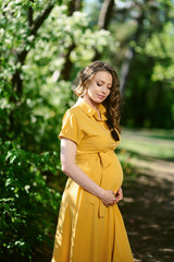 Attractive young pregnant woman in a long yellow dress outside. Warm sunny summer weather. Between the second and third trimesters of pregnancy