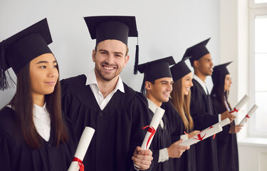 Happy student man with diploma in hands looking at camera standing next to his multiethnic classmates friends. Group of people in graduation gowns standing in a row near the wall in the classroom.