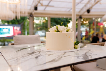 Soft focused shot of beautiful white wedding or birthday cake with roses flowers on marble table, restaurant background. Festive event sweet bakery