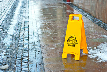 Yellow danger sign. Symbol of a slippery sidewalk. Inscription attention in different languages. Plate near the cobblestones in the spring.
