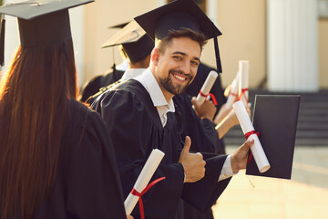 Fototapeta Portrait of a graduate student with the diploma and in the black academic gown raises his thumbs up at graduation at university. Student looks at the camera and smiles sincerely. obraz