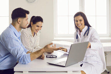 Obraz premium Expecting a baby. Smiling nurse showing scan picture to future parents. Happy couple, husband and his pregnant wife, looking at ultrasound image on computer screen sitting at table at doctor's office