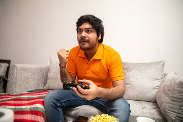 Asian Indian man gaming, streaming and watching TV alone in living room on a grey sofa and white table