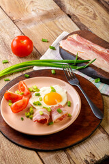 Scrambled eggs with bacon with tomato and green onion and on a wooden table in a plate with a fork next to chopped bacon and tomatoes.