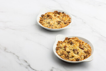 Two plates of delicious apple crumble pie isolated on marble background. Close-up of double crumble pies with fruits, nuts and cinnamon in white bowls. Dessert menu concept. Copy space.
