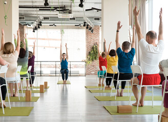 group of senior people doing yoga with chairs and with attractive young male teacher indoor