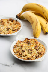 Two plates of delicious apple crumble pie isolated on marble background with bananas by side. Close-up of double crumble pies with fruits, nuts and cinnamon in white bowls. Dessert menu concept.