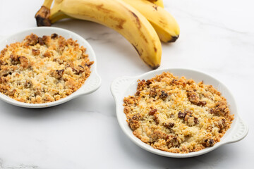 Two plates of delicious apple crumble pie isolated on marble background with bananas by side. Close-up of double crumble pies with fruits, nuts and cinnamon in white bowls. Dessert menu concept.