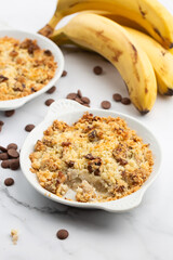 Two plates of delicious apple crumble pie isolated on marble background with bananas and choco dots on it. Close-up of double crumble pies with fruits and nuts in white bowls. Dessert menu concept.