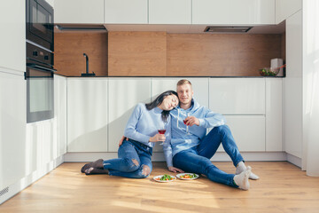 Couple in love eating a salad of vegetables, sitting on the floor in the kitchen, and drinking wine
