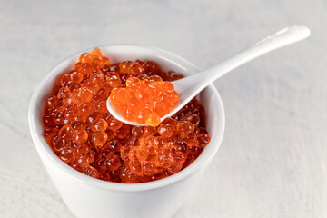 Close up delicious, crumbly, large red caviar in a white cup with a spoon on a white background.