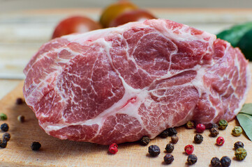 A piece of raw meat with peppercorns and bay leaves on a cutting board. In the background fresh vegetables - tomatoes and cucumbers.