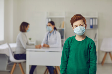 Fototapeta na wymiar Portrait of little boy wearing face mask. Healthy child in medical mask standing against blurred doctor's office and looking at camera. Concept of healthcare for children and prevention of infection