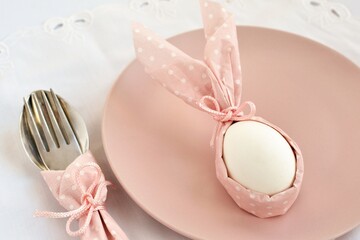Decorative serving  of Easter table. A white egg with rabbit ears from a pink napkin on a plate with a spoon and fork.