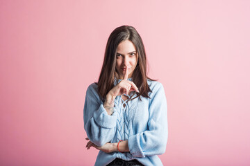 Portrait of a brunette girl on a pink background who holds a finger to her mouth