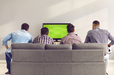 Back view of young men sitting on comfortable grey sofa and watching live football game on TV. Group of 4 friends relaxing on soft couch at home and enjoying interesting soccer match on television