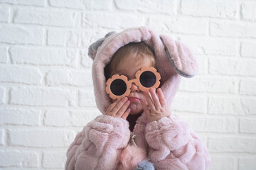 A child in a bunny costume and sunglasses makes faces against a white brick wall background.