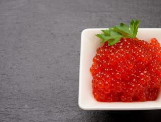 fresh grained red chum salmon caviar in a white ceramic bowl on a black slate stone background