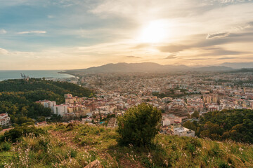 Fototapeta na wymiar Beautiful panoramic sunset view of the city of Malaga from a viewpoint on a hill with lawn and plants, overlooking the city, the sea, the mountains and the cloudy sky in the background.