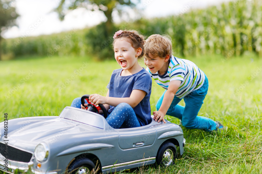 Wall mural Two happy children playing with big old toy car in summer garden, outdoors. Boy driving car with little girl inside. Laughing and smiling kids. Family, childhood, lifestyle concept