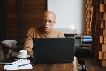 Confident prosperous African-American young top-manager wearing formal shirt having coffee and doing paperwork, working on generic laptop, using free wireless internet connection at restaurant