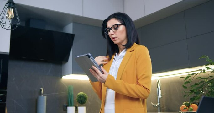 Thoughtful likable concentrated adult black-haired woman in glasses and stylish orange jacket standing in cozy kitchen and using tablet device