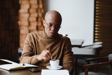 Concentrated black male entrepreneur reads paper documents sitting in cafe with coffee near window.