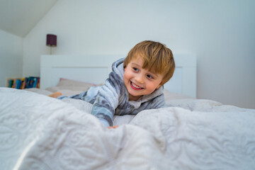 Fototapeta premium Little blond toddler boy relaxing in bed. Home leisure. Laughing child playing in bed.