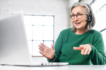 Active mature woman using a laptop for remote work from the home office. Smiling elderly lady...