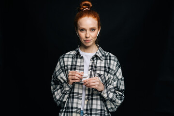 Beautiful red-haired young woman wearing wireless earphones looking at camera on isolated black background. Pretty redhead lady model emotionally showing facial expressions, copy space.