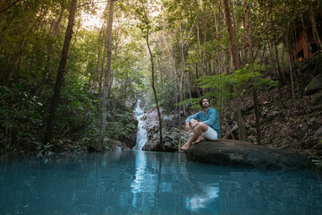 A man sits on a stone in a lake in a tropical forest.