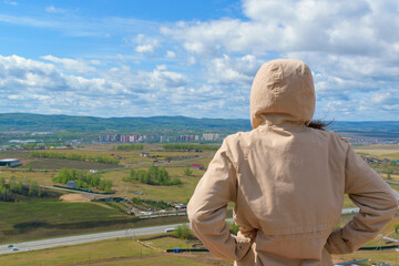 A woman in a gray raincoat looks down at the spring landscape. Back view, hood. Road, green trees, mountains, houses. Blue sky with clouds.