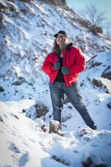 A beautiful girl in a red down jacket, jeans and a black baseball cap stands in the middle of the snow dunes somewhere in the mountains and looks into the distance.