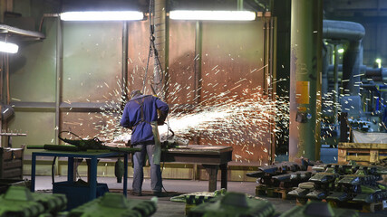 Workers in protective equipment in a foundry work on a casting with a grinding machine at the...