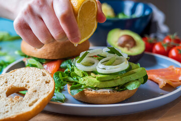 Bagel sandwiches in the making. Gluten free toasted bagel with avocado, mayo, lettuce, cucumber and onion. Hand squeezes lemon on top and salmon bagel background. Healthy breakfast, fun vibrant colors