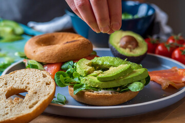 Bagel sandwiches in the making. Gluten free toasted bagel with garlic mayo, avocado lettuce and cucumber. Hand adding salt and salmon bagel in background. Healthy breakfast with fun vibrant colors