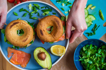 Hands making bagel sandwiches. Gluten free toasted bagels with smoked salmon and cream cheese and with avocado, cucumber, lettuce, and onions. Homemade Healthy breakfast, fun vibrant colors. Top view