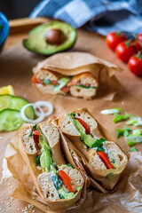 Stuffed toasted bagel sandwich cut into half with avocado, mayo, cucumber, lettuce and tomatoes. Salmon bagel in the background, wrapped with paper. Gluten free healthy breakfast, vibrant fun colors 