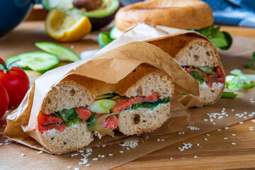 Stuffed toasted bagel sandwich cut into half with smoked salmon, cream cheese, cucumber lettuce. Avocado bagel in the background on a baking sheet. Gluten free healthy breakfast, vibrant fun colors 