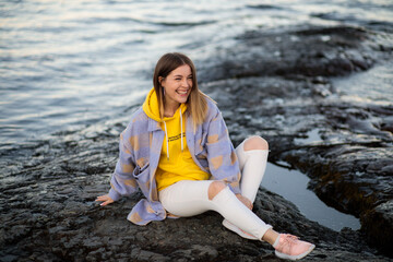 A beautiful girl sits on a rocky shore and smiles. A stylish beauty in autumn clothes sits on a stone by a pond and laughs with happiness and good mood.

