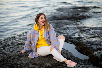 A beautiful girl sits on a rocky shore and smiles. A stylish beauty in autumn clothes sits on a stone by a pond and laughs with happiness and good mood.
