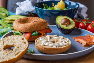 Bagel sandwiches in the making. Gluten free toasted bagel with garlic mayo ready to add avocado lettuce, cucumber and onions. Smoked salmon bagel in background. Healthy breakfast, fun vibrant colors