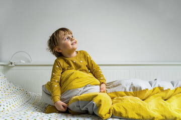 Little toddler boy is sitting in bed. Child waking up in the morning with smile. Happy kid resting...